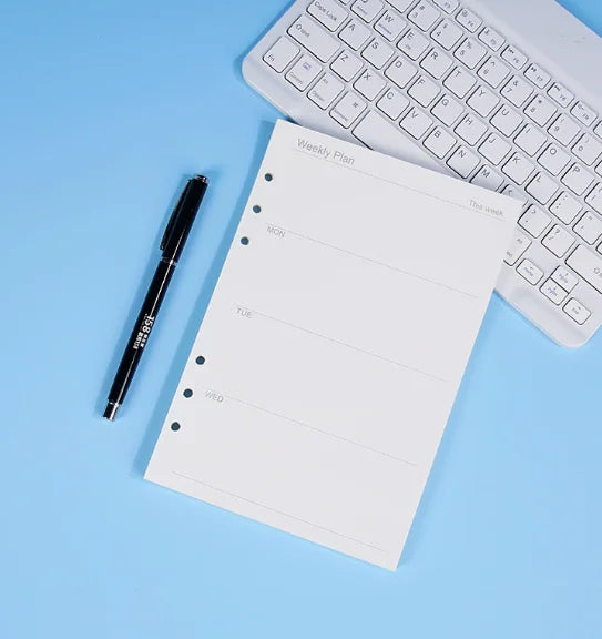 Una página de cuaderno de planificación semanal junto a un bolígrafo negro y un teclado de laptop blanco sobre un fondo azul 