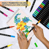 A hand holds a yellow brush marker drawing flowers on a notebook surrounded by a large set of vibrant dual-tip pens.