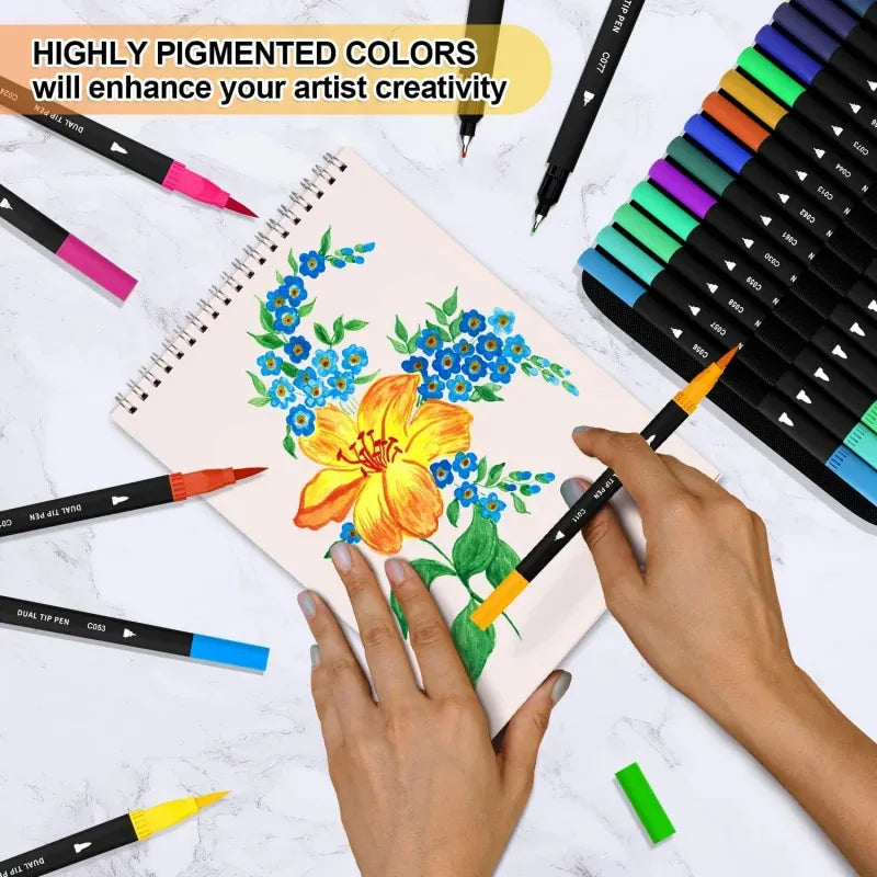 A hand holds a yellow brush marker drawing flowers on a notebook surrounded by a large set of vibrant dual-tip pens.