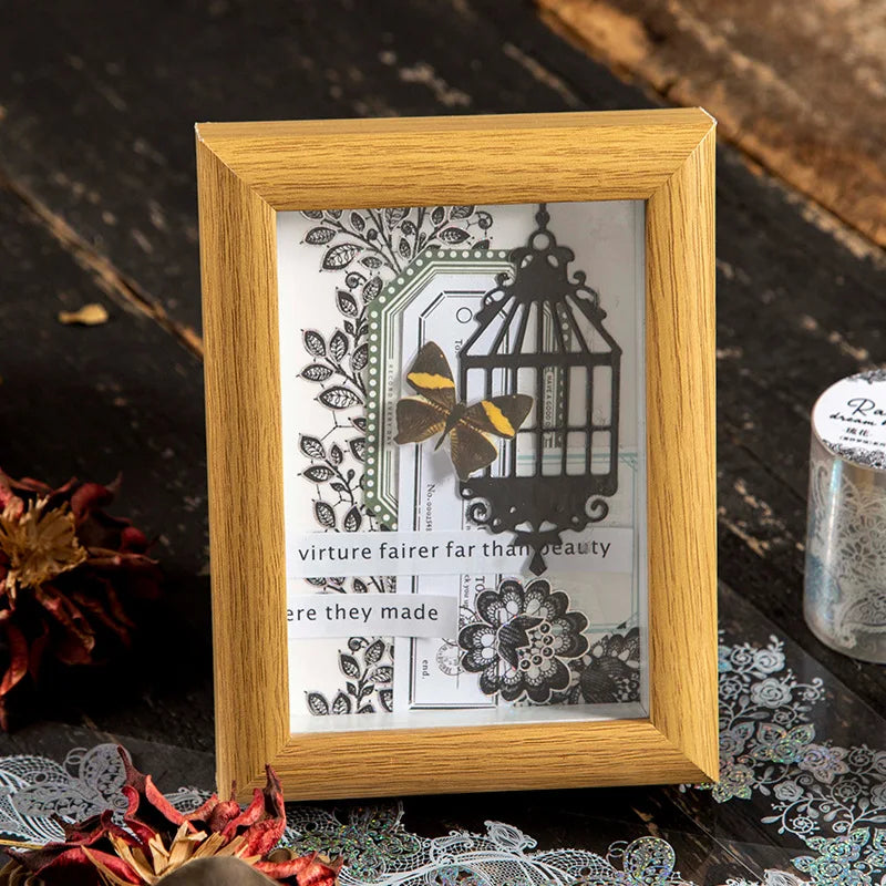 A wooden frame holds a decorative silver stamping lace tape featuring botanical designs and a birdcage silhouette.