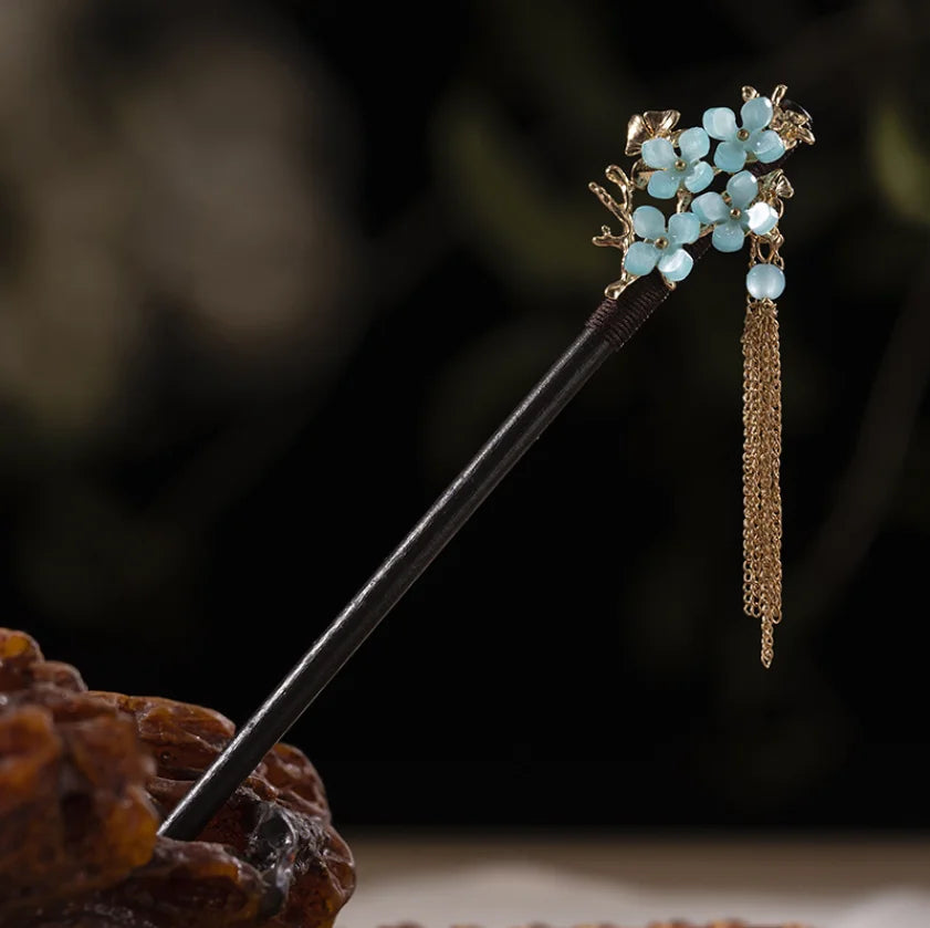 Une épingle à cheveux dorée avec des accents floraux bleu clair et une pompon doré, sur fond sombre.