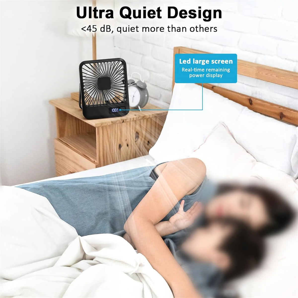 A black portable USB rechargeable desk fan is shown next to a wooden nightstand in a bedroom setting.