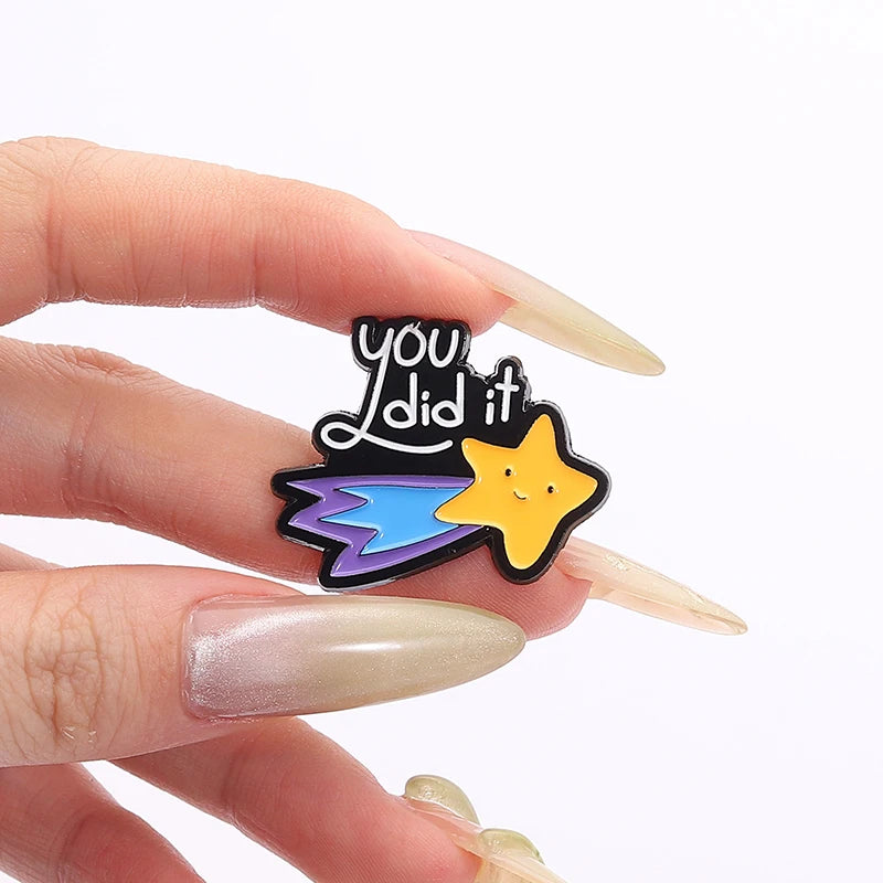 A hand holds a black enamel pin featuring the text you did it and a yellow star with blue accents.