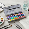 An open watercolor paint set box displaying a palette of solid colored paints and various brushes on a light wooden surface.