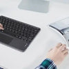 A person is using a black wireless keyboard and touchpad on a white desk while looking at an open magazine.