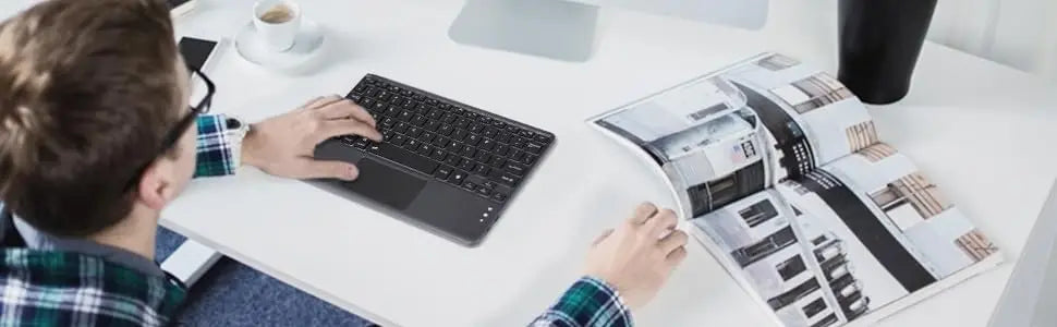 A person is using a black wireless keyboard and touchpad on a white desk while looking at an open magazine.