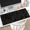 A black gaming mouse pad featuring white topographic line patterns placed on a desk next to a computer monitor.