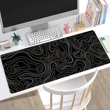 A black gaming mouse pad featuring white topographic line patterns placed on a desk next to a computer monitor.