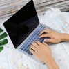 Hands typing on a silver silicone keyboard cover for a MacBook Air laptop on a light wooden surface.
