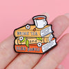 An enamel lapel pin depicting a stack of books, a teacup, and leaves with the text Drink Tea and Read Books.