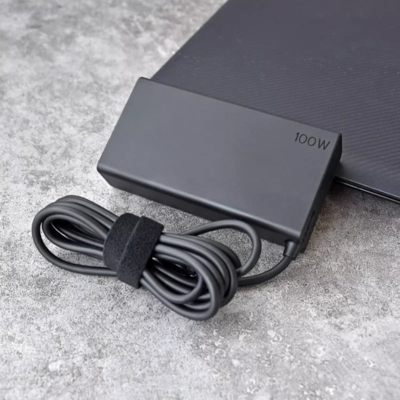 A black 100W USB-C PD laptop charger adapter with a coiled cable lying on a gray surface.