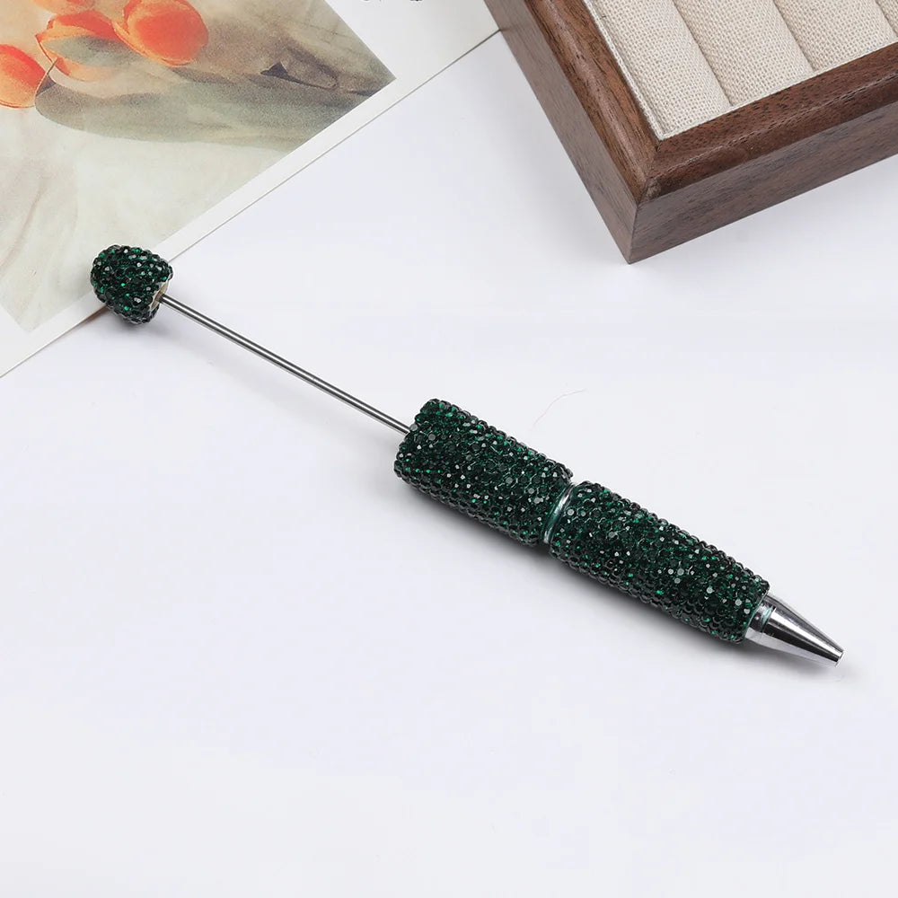 A dark green rhinestone beaded pen accessory with a silver tip is displayed on a white surface.