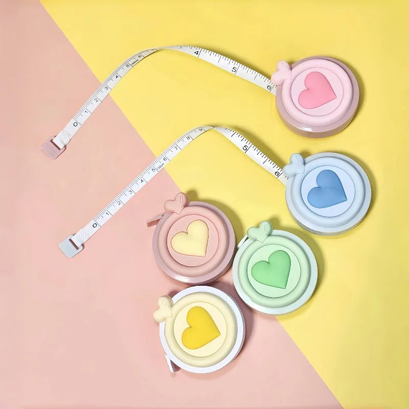 Cinta métrica en forma de corazón rosa y amarillo con pegatinas de corazón de varios colores sobre un fondo rosa claro.