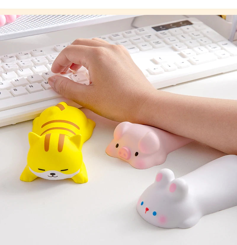 A hand rests on a white keyboard with a yellow cat and a pink pig shaped ergonomic wrist rest nearby.