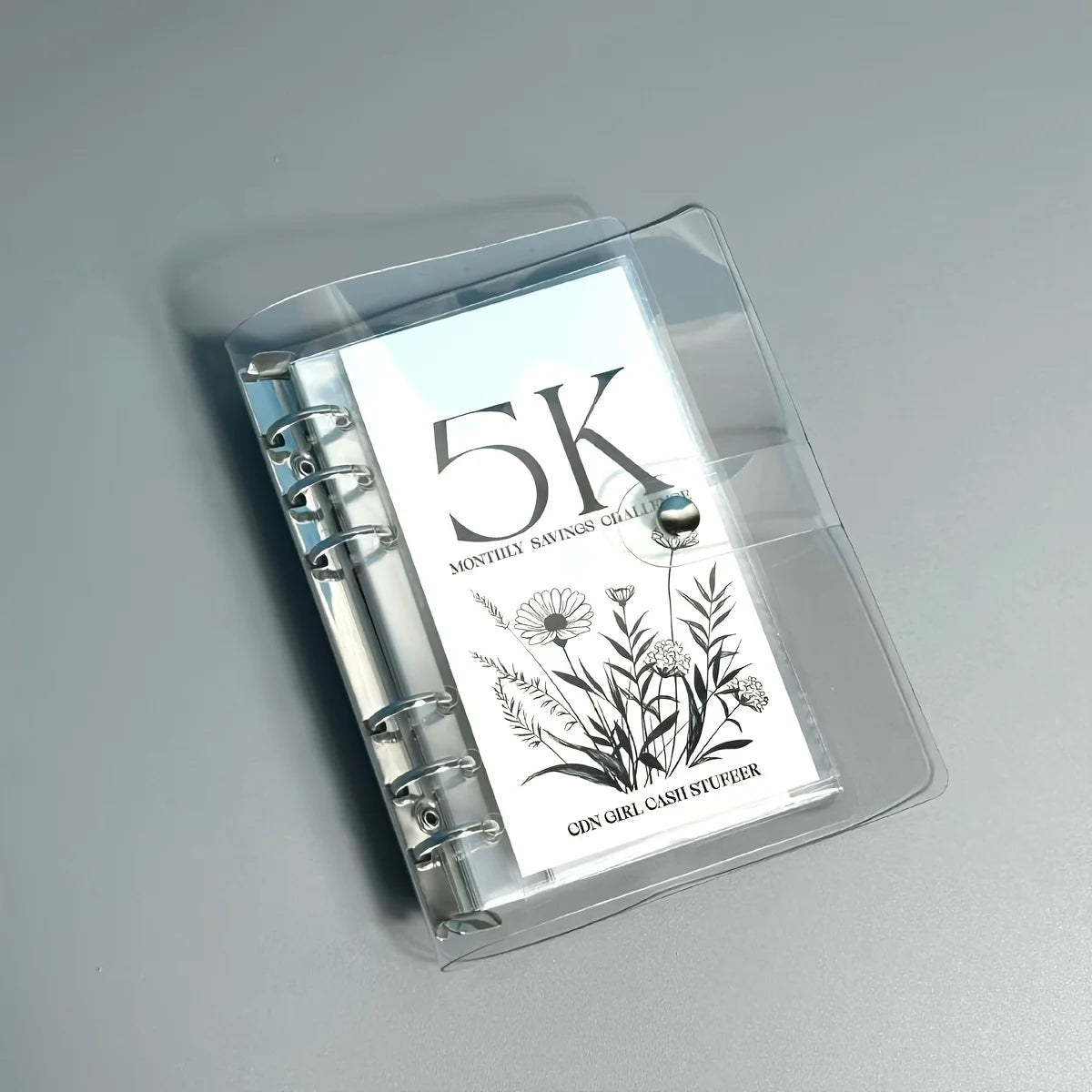 A clear plastic binder for a 5K monthly savings challenge featuring floral illustrations.