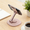 A pink foldable desktop phone stand holding a smartphone, placed on a wooden table next to a notebook and a cup.