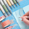 A hand is using a silver retractable craft knife pen set to cut blue paper on a grid.