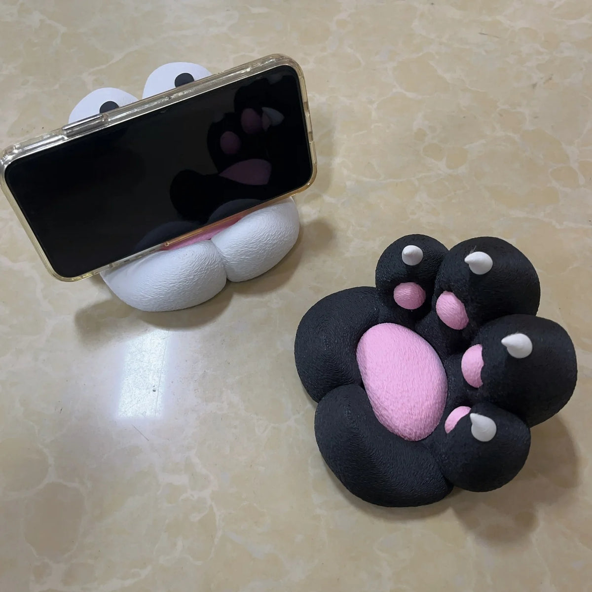 A black phone is resting on a white, 3D printed cat paw shaped stand with anti-slip features.