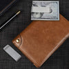 A brown leather money clip wallet with a silver card and a gold pen resting on a dark surface.