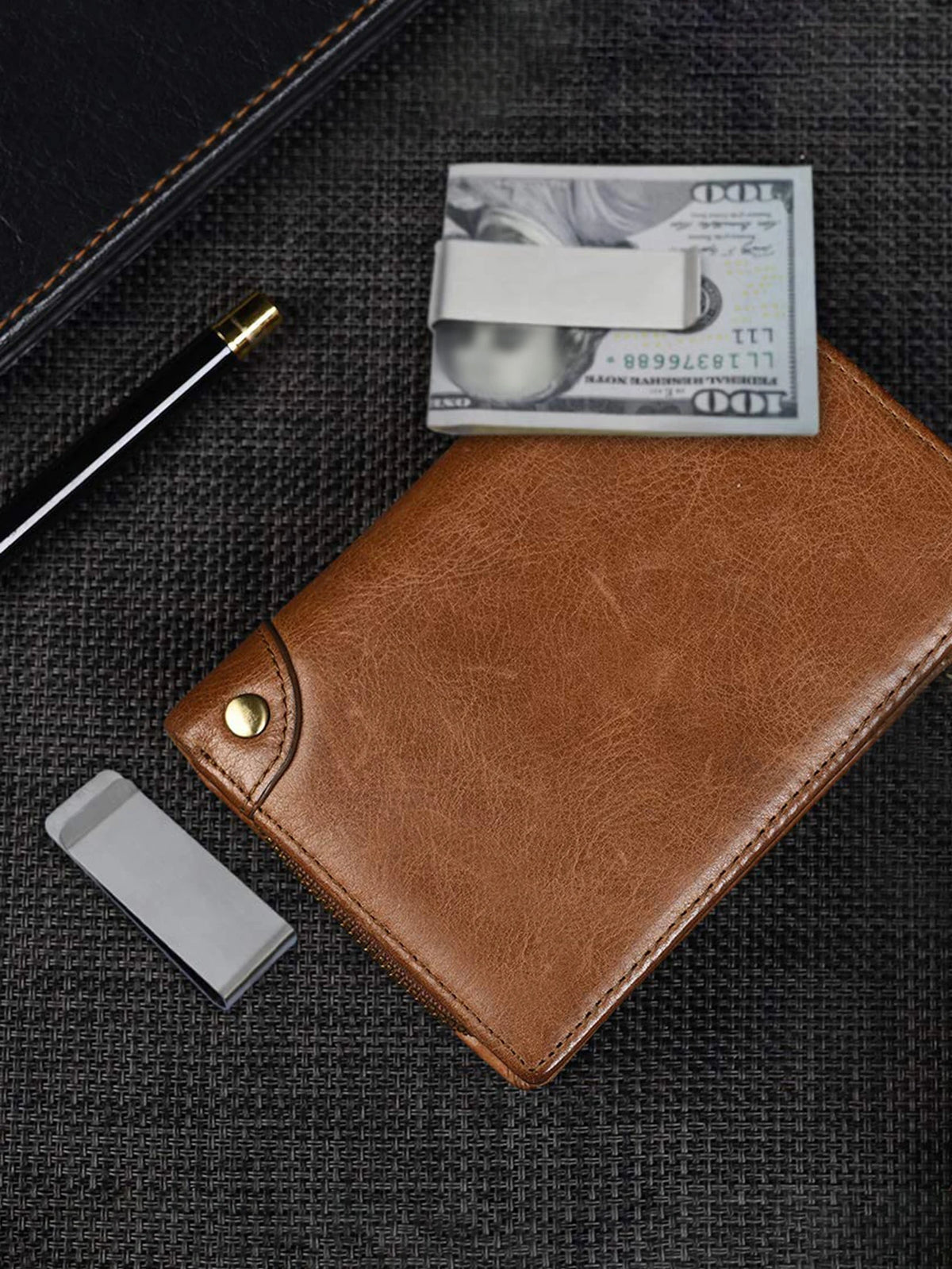 A brown leather money clip wallet with a silver card and a gold pen resting on a dark surface.