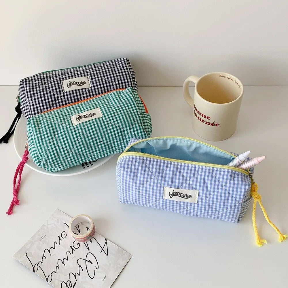 A collection of three fabric pencil cases in green, blue, and purple checkered patterns are displayed next to a white mug and