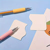 A set of retractable craft knife pens, including a yellow and a pink pen, resting on light blue paper.