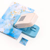 A light blue embossing punch machine with a beige handle and a sheet of blue watercolor paper featuring gold accents.