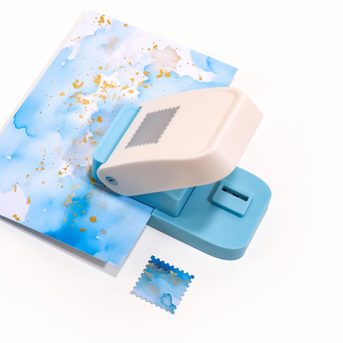 A light blue embossing punch machine with a beige handle and a sheet of blue watercolor paper featuring gold accents.