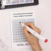 A hand is writing on a tracker sheet for a 100 envelope saving challenge, next to a stack of cash