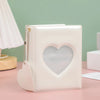 A white hollow photo card binder with a heart-shaped cutout and a silver chain detail resting on a pink surface.