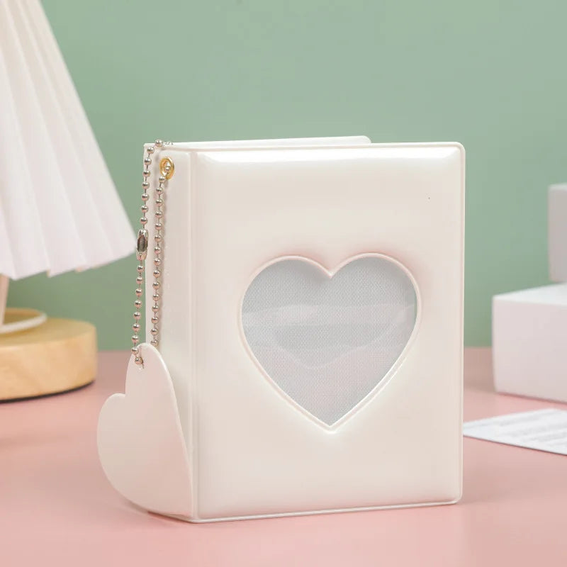 A white hollow photo card binder with a heart-shaped cutout and a silver chain detail resting on a pink surface.