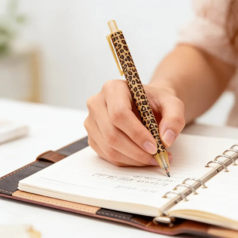 A hand is writing in a leather-bound notebook with a leopard print retractable ballpoint pen.
