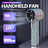A sparkling diamond-like handheld fan with a LED display and rechargeable battery is shown on a dark surface.