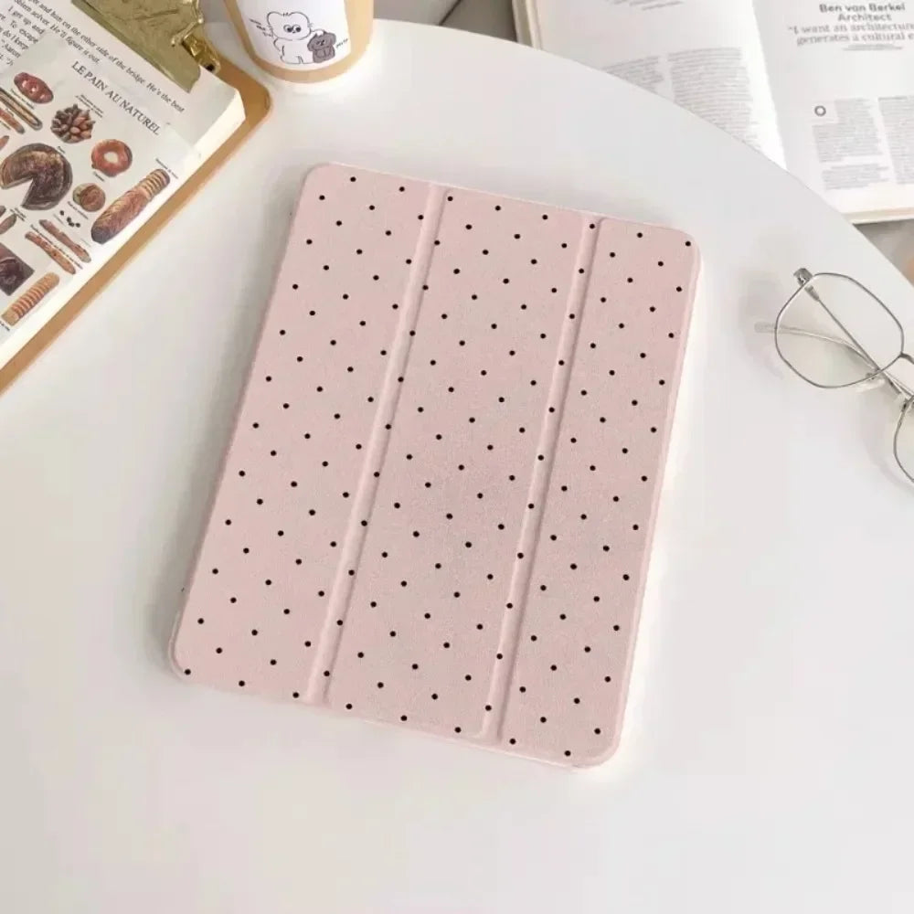 A pink acrylic protective skin with small black dots is displayed on a white table next to an iPad case and eyeglasses.