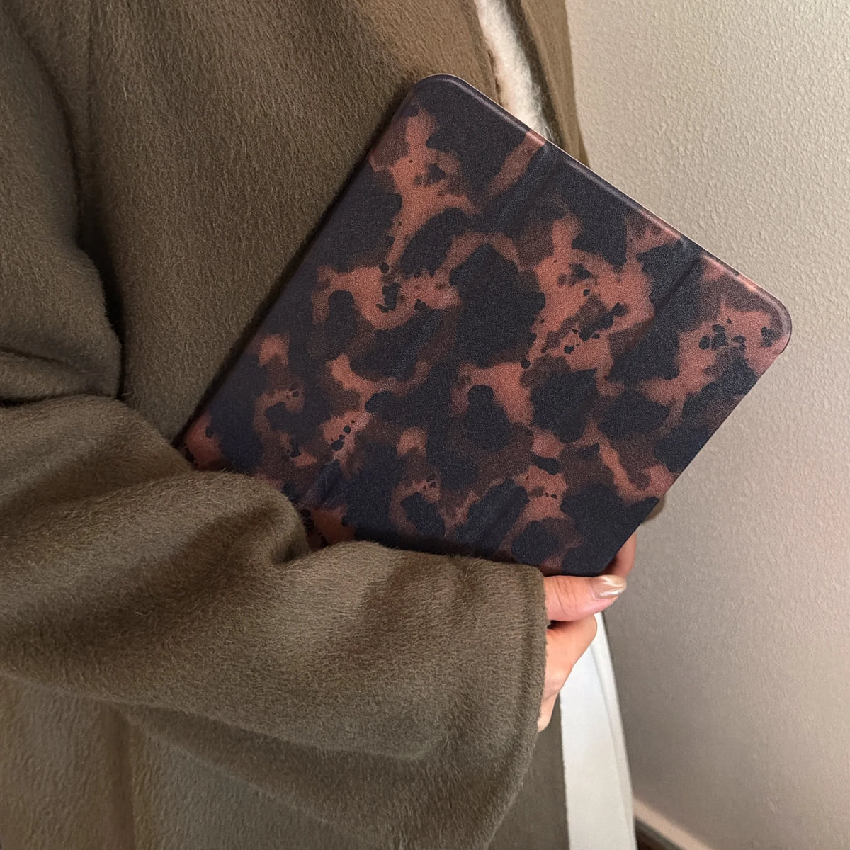 Dunkelbraunes Leopardenmuster-Leder-Tablet-Cover wird in der Hand einer Person gegen einen neutralen Mantel gehalten.