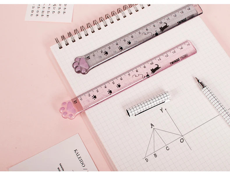 A pink plastic straight ruler marked with cat paw shapes is shown next to graph paper and a pencil.