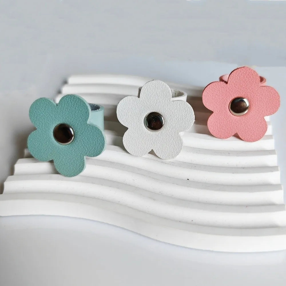 Three leather flower cable organizers in mint green, white, and pink are displayed on a white wavy surface.