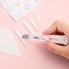 A hand holds a pink portable pet claw tool knife with a blade, positioned near white paper scraps on a pink background.
