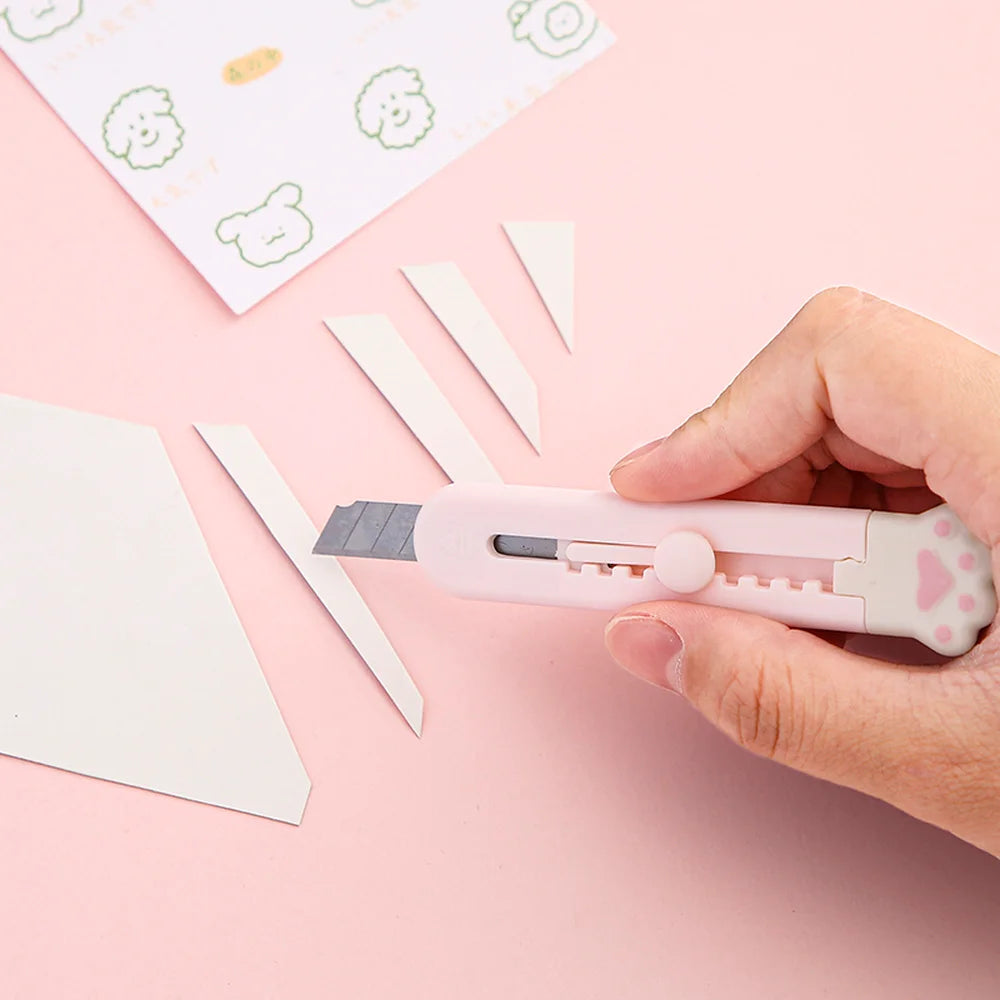 A hand holds a pink portable pet claw tool knife with a blade, positioned near white paper scraps on a pink background.