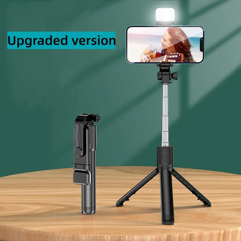 A black selfie stick tripod with a fill light and wireless remote is displayed on a wooden table.