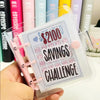 A hand holds a clear binder titled Savings Challenge with dollar amounts and text on the cover.