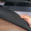 A hand holds a black overlocked mouse pad on a wooden desk in front of an open laptop.