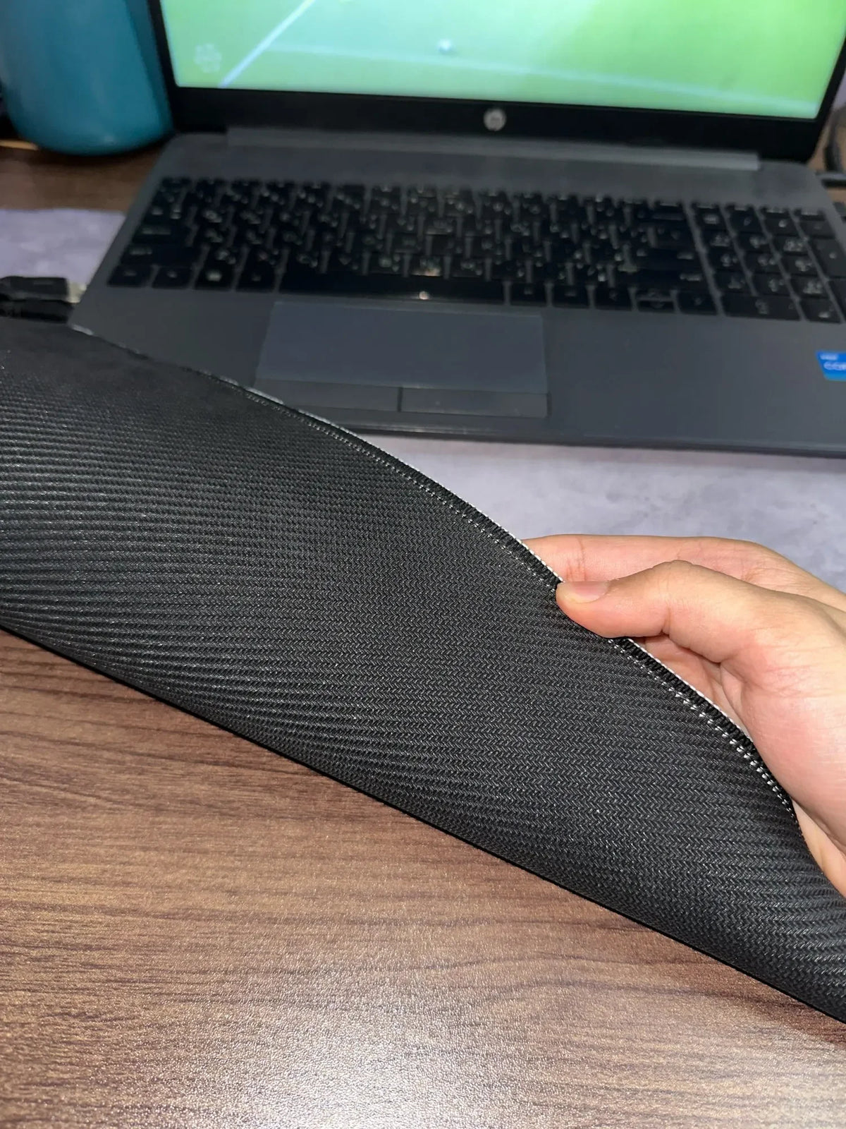 A hand holds a black overlocked mouse pad on a wooden desk in front of an open laptop.