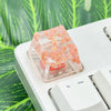 A small clear plastic container filled with translucent pink and orange botanical resin keycaps resting on a white mechanical