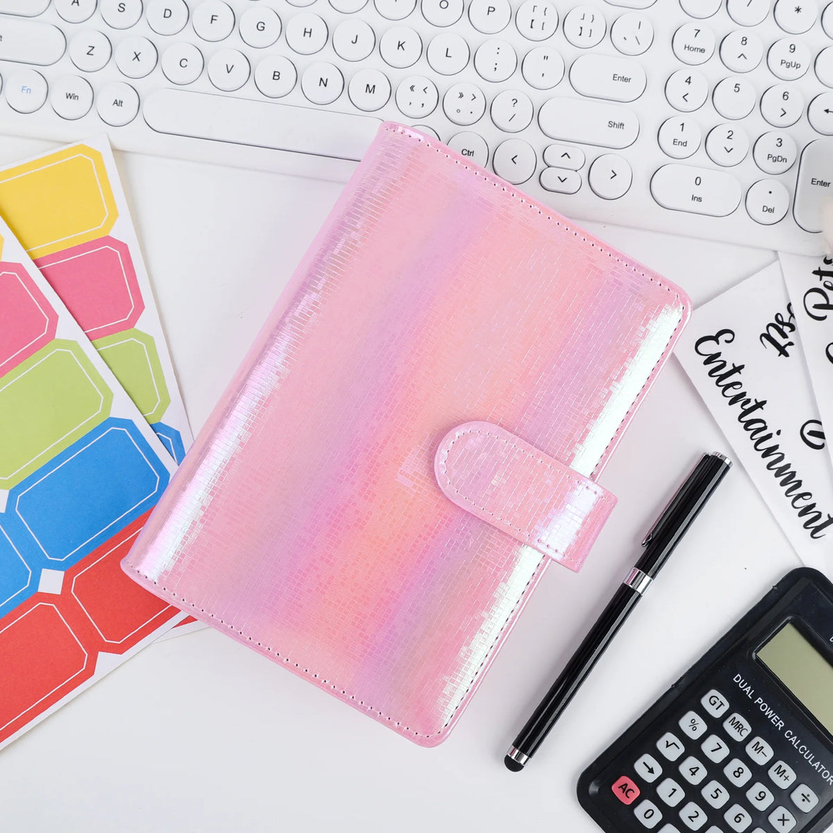 Un cahier rose pailleté et une calculatrice noire sont montrés sur un bureau blanc à côté d'un clavier.