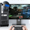 Eine Hand hält eine schwarze Gaming-Konsole vor einem Computerset mit PC-Gehäuse und Monitor.