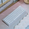 A white mechanical keyboard with translucent keycaps is displayed on a pink surface next to a book.