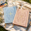 A light blue A5 dot grid notebook with a gold foil botanical cover resting on a linen cloth.