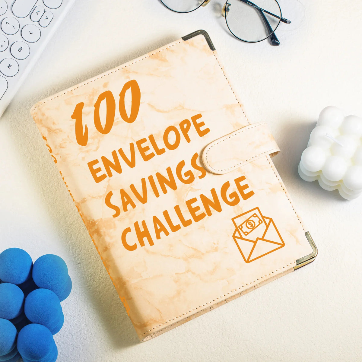 A beige marble budget binder titled 100 Envelope Savings Challenge rests on a white surface with blue foam balls nearby.