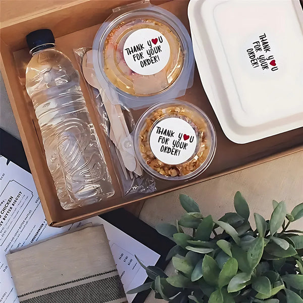 A wooden box contains several clear plastic containers with thank you stickers, some containing food items and a bottle of li